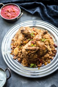 Chicken on a bed of spiced kabsa rice with mixed nuts and raisins, with two small bowls of tomato sauce and yogurt on the side.