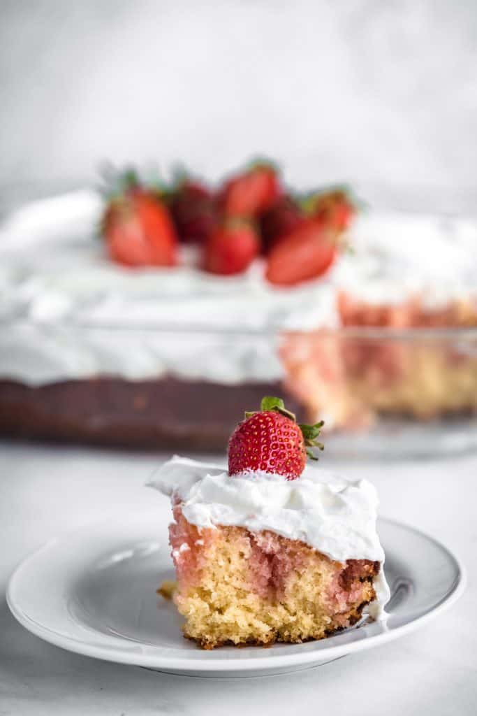 A slice of strawberry poke cake on a plate in front of the remainder of the cake