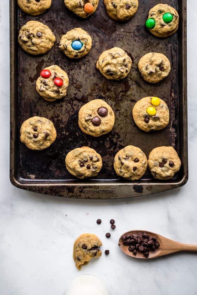 Distressed baking sheet with mini chocolate chip cookies cooling on it and a wooden spoon with chocolate chips on it