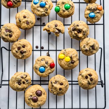 A black wire cooling rack with mini chocolate chip cookies on it, some with m&m's and a glass of milk at the bottom of the photo