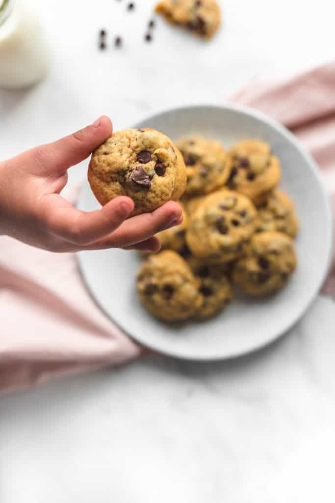 A child hand grabbing a mini chocolate chip cookie off a plate that's on a pale pink kitchen towel