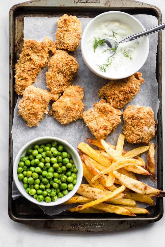 Parchment paper lined baking tray with baked crispy fish and chips on it along with a bowl of peas and homemade tartar sauce