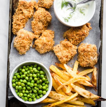 Parchment paper lined baking tray with baked crispy fish and chips on it along with a bowl of peas and homemade tartar sauce