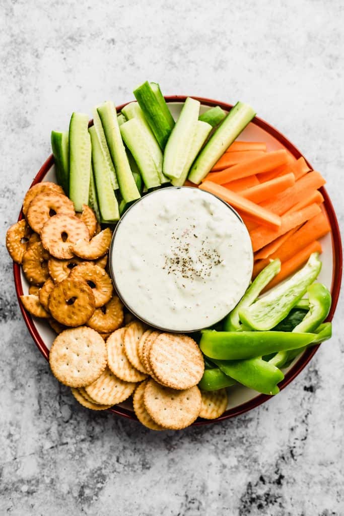 Plate with cut up carrots and cucumbers, crackers and pretzels with labneh dip in a bowl in the middle