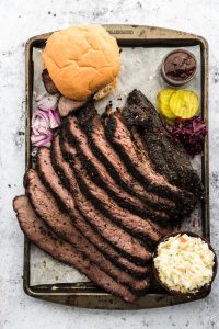 Sliced cooked sous vide brisket on a tray with bread buns, pickles and barbecue sauce