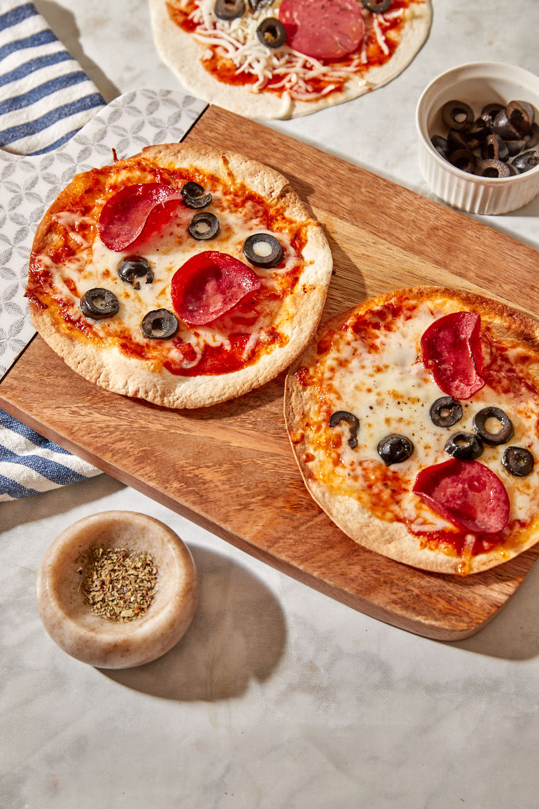 two air fryer tortilla pizzas with pepperoni and sliced black olives on a wooden cutting board.