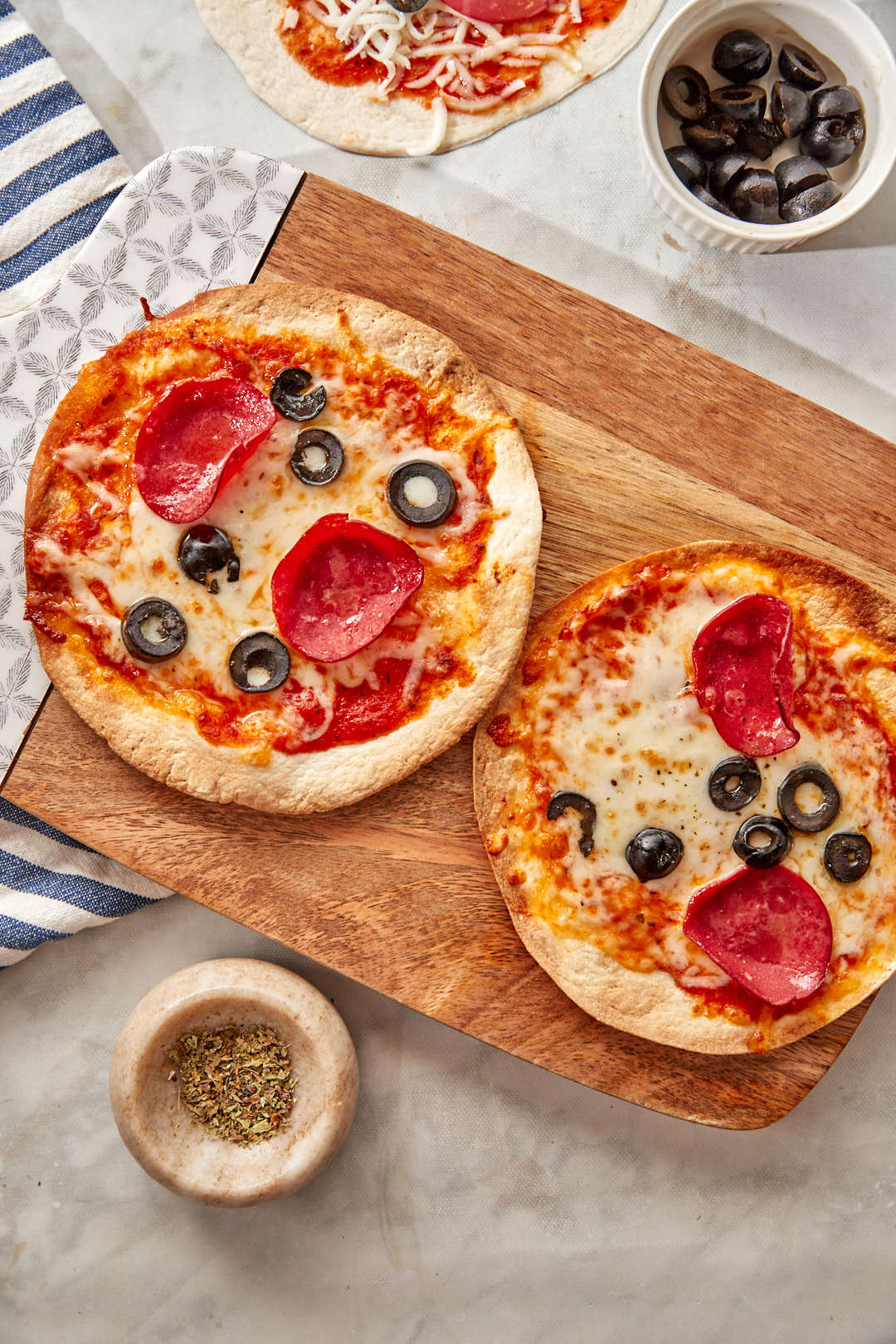 two air fryer tortilla pizzas with pepperoni and sliced black olives on a wooden cutting board.