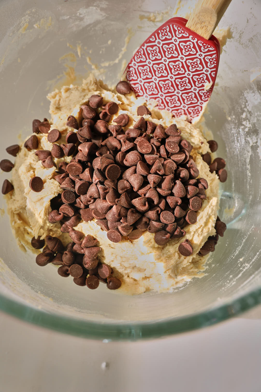chocolate chips added to cookie dough with a red spatula in a glass bowl.