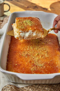 a slice of cream stuffed basbousa being lifted out of a white pan wih a gold cake server.