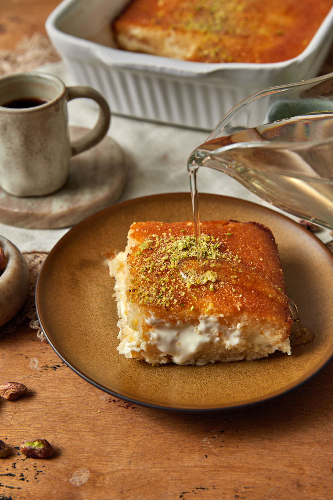 sugar syrup being poured onto a slice of basbousa with cream on a brown plate with Arabic coffee on the side.