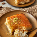 a slice of basbousa with a fork taking a bite out, with another slice on a plate in the background.