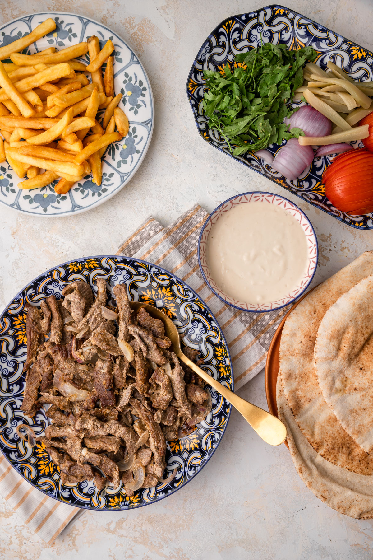 Beef shawarma on a plate with other plates of fries, tahini, vegetables.