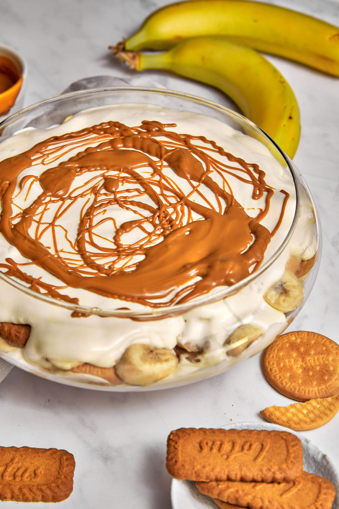 a big serving dishf ull of banana pudding with bananas in the background, and biscoff biscuits on the table.