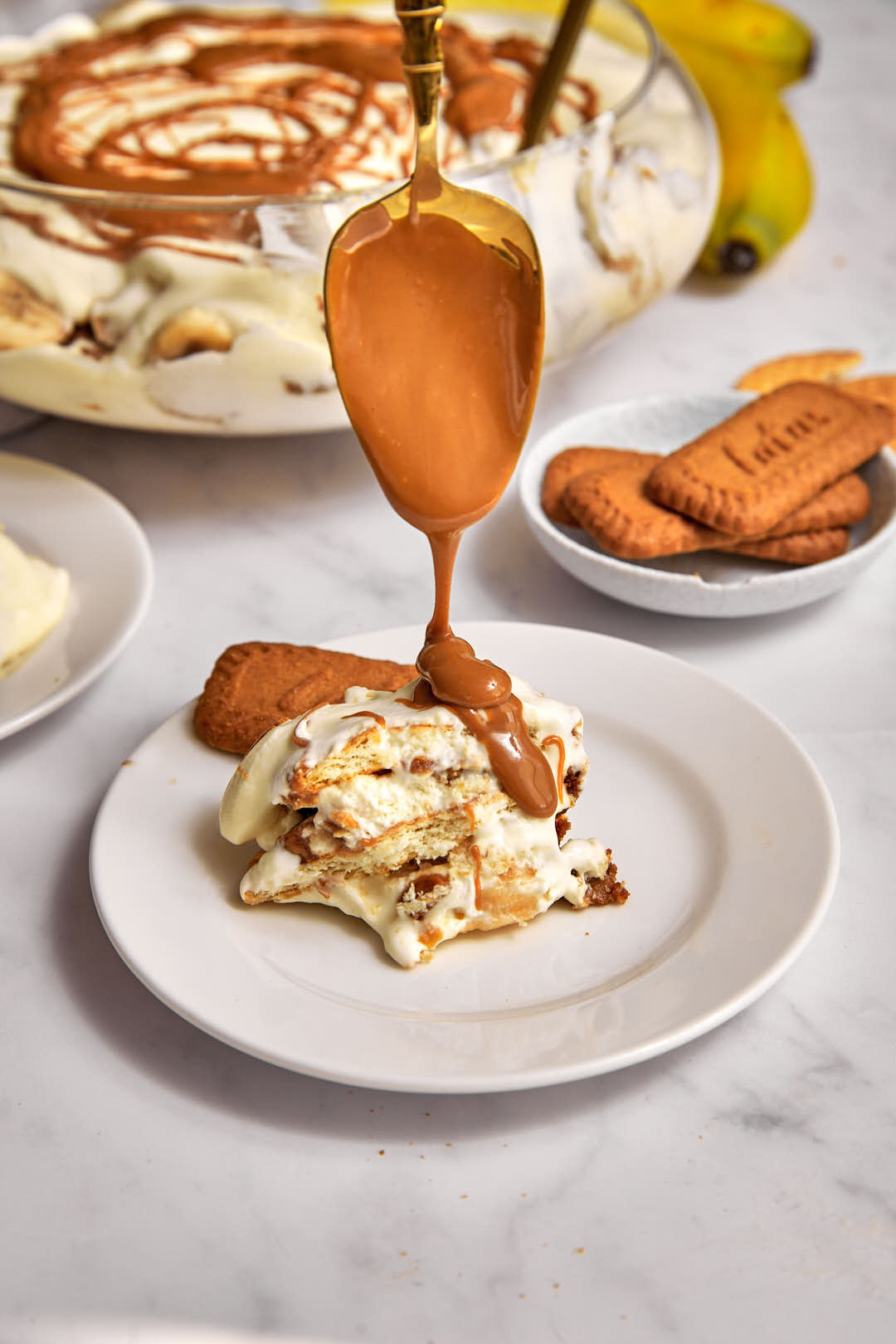 biscoff spread being drizzled over a scoop of banana biscoff pudding on a white plate.