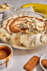 a scoop of biscoff banana pudding being lifted out of a glass trifle bowl.