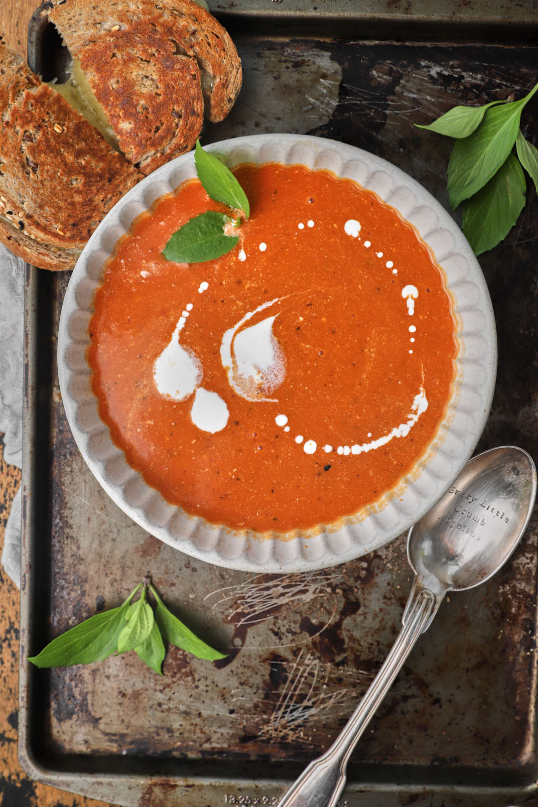 a bowl of tomato soup on a baking tray with a spoon, spring of basil, and half a grilled cheese sandwich.