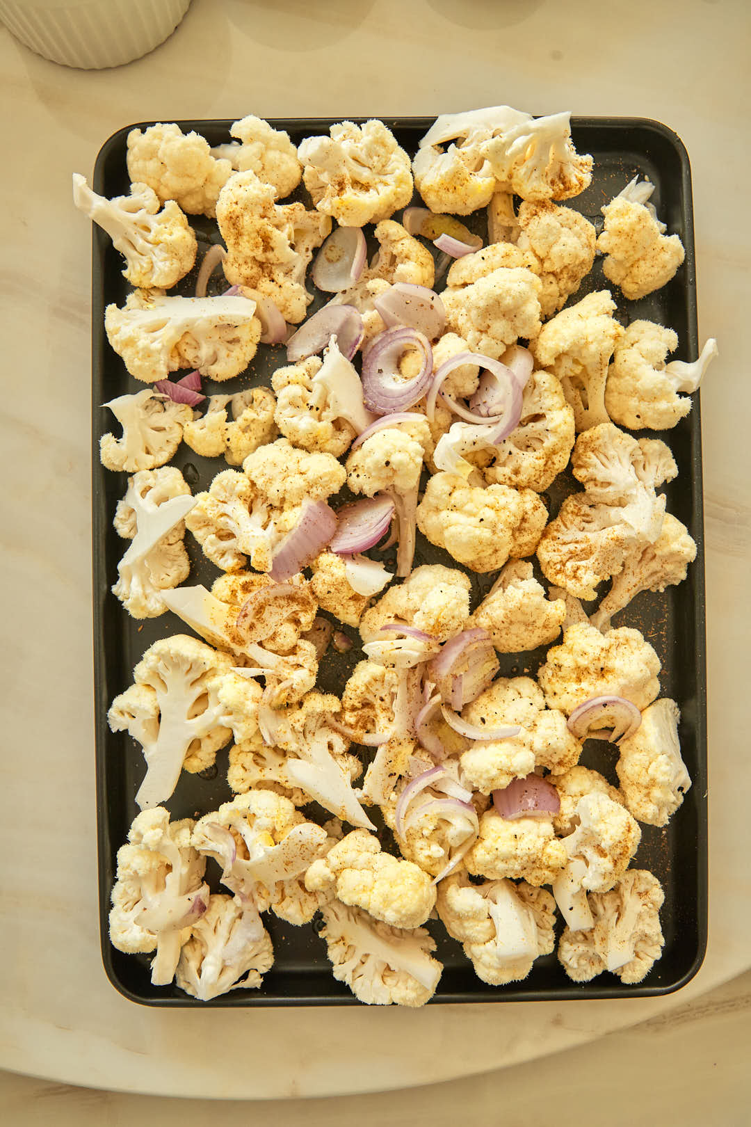 Raw cauliflower florets and sliced red onion seasoned on a baking sheet before roasting