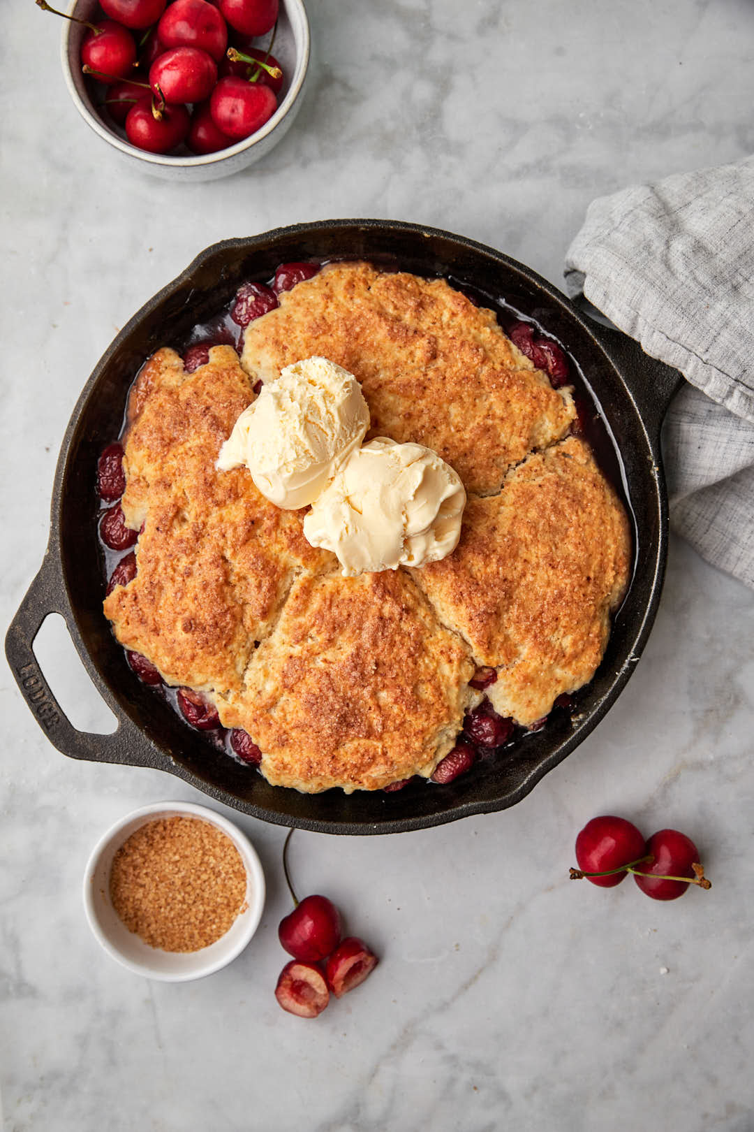 Cast iron skillet cherry cobbler on white surface with fresh cherries in the background.
