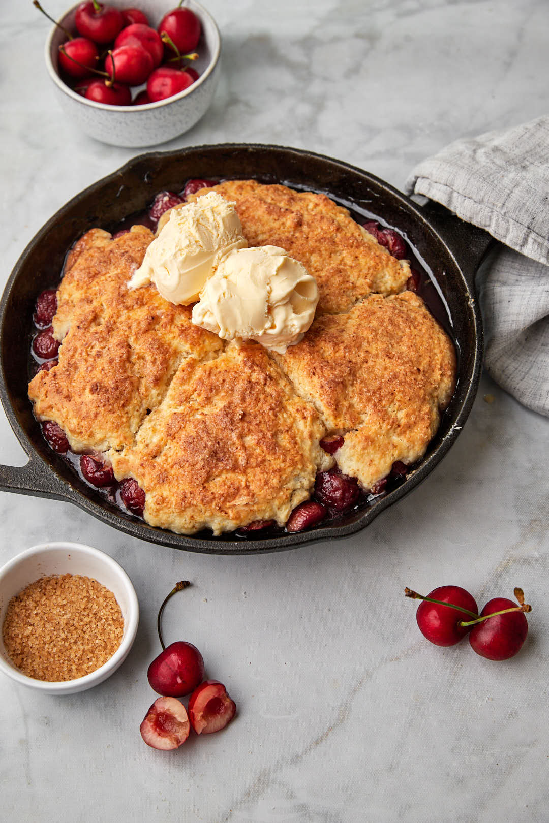 Cast iron skillet with cherry cobbler with a kitchen towel wrapped around the handle and cherries in the backdrop.