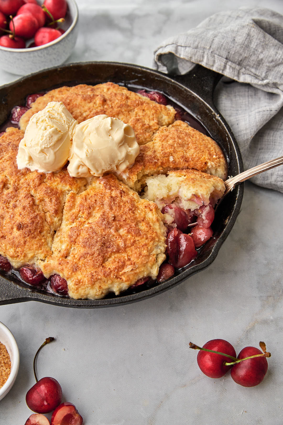 A scoop being taken out of a cherry cobbler with biscuits with cherries in the background.