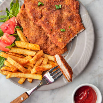 two pieces of chicken escalope on a plate with a bite being taken out with a fork and french fries and a salad on the plate.