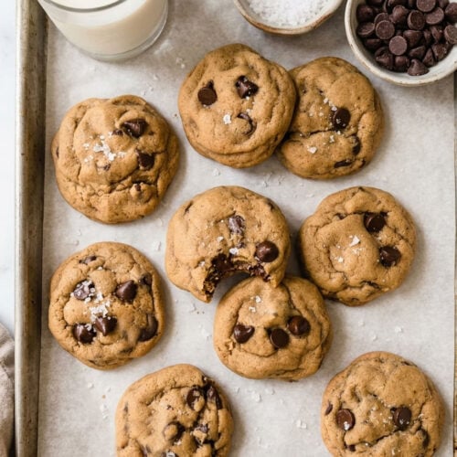 baking tray with chocolate chip cookies, a small bowl of chocolate chips, a small bowl of sea salt and a glass of milk.