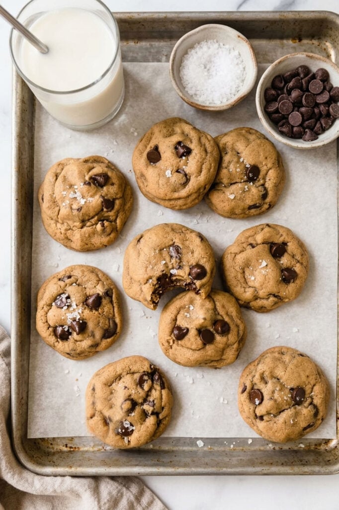 baking tray with chocolate chip cookies, a small bowl of chocolate chips, a small bowl of sea salt and a glass of milk.