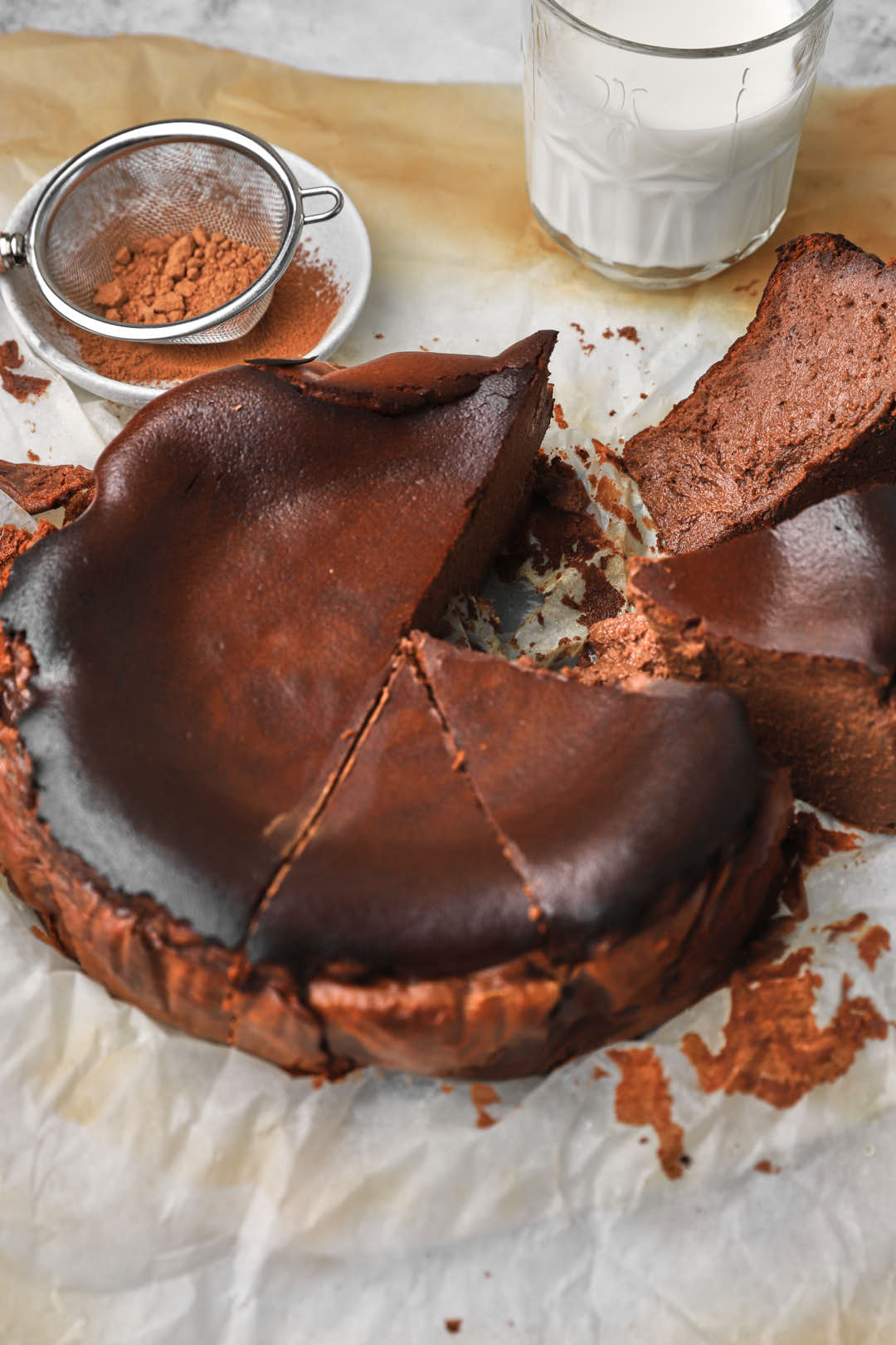 a whole chocolate basque cheesecake with a slice being taken out and some cocoa powder and milk in the background.