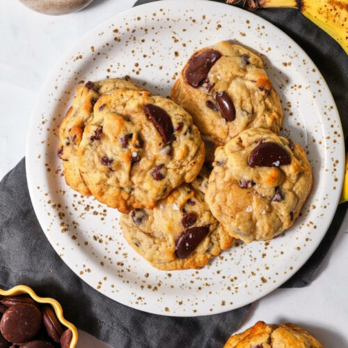 a plate of chocolate chip banana bread cookies on a dark kitchen linen with some bananas, sea salt, and chocolate, in the backdrop.