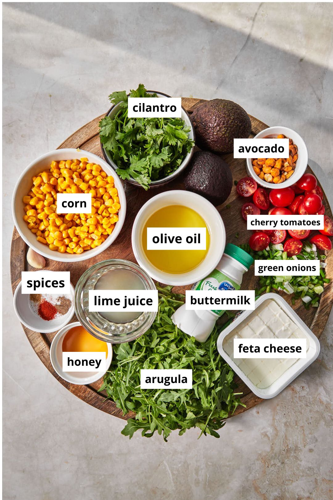 ingredients needed to make a corn avocado and arugula salad on a circular wooden platter.