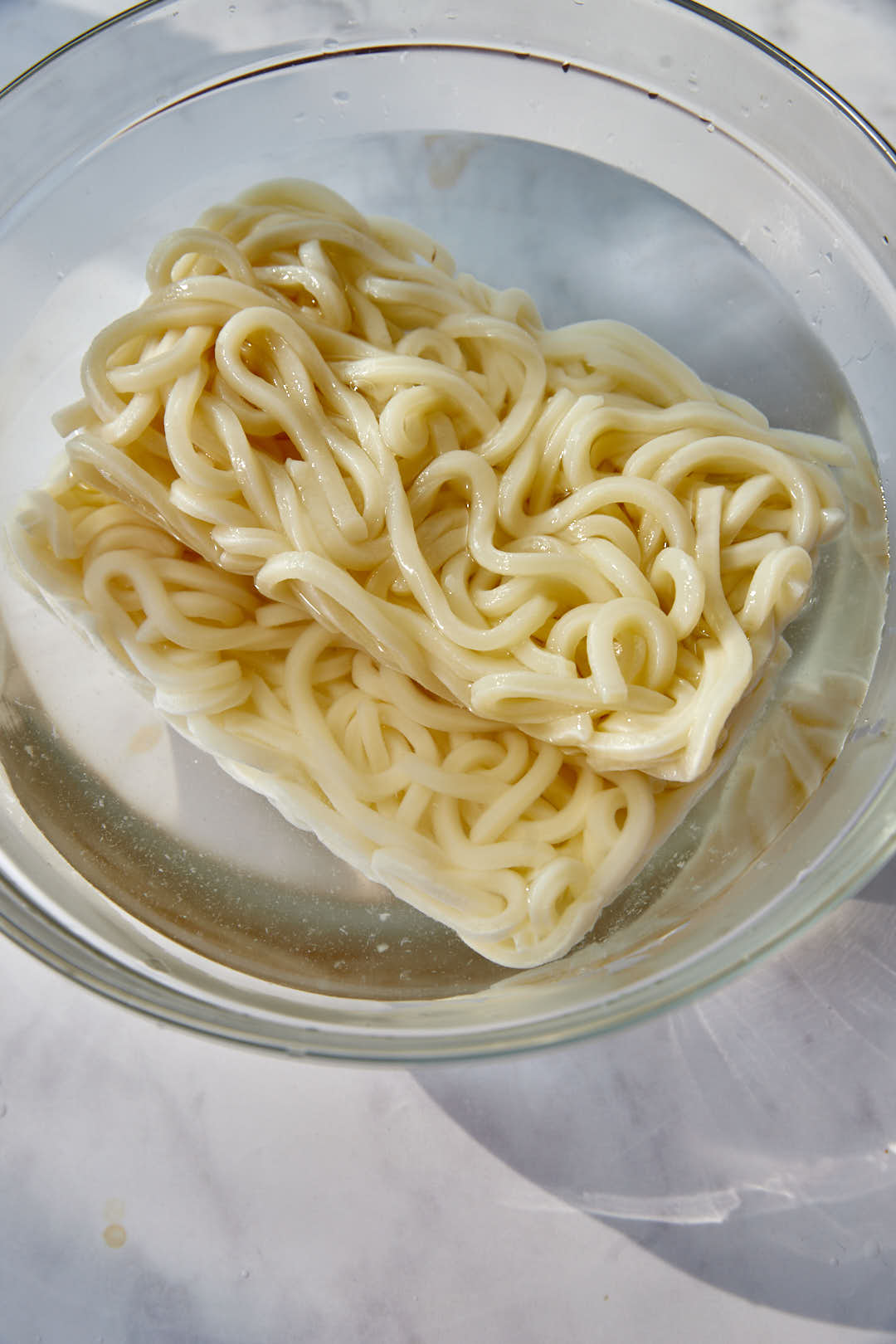 Udon noodles soaking in boiling water in a glass bowl.