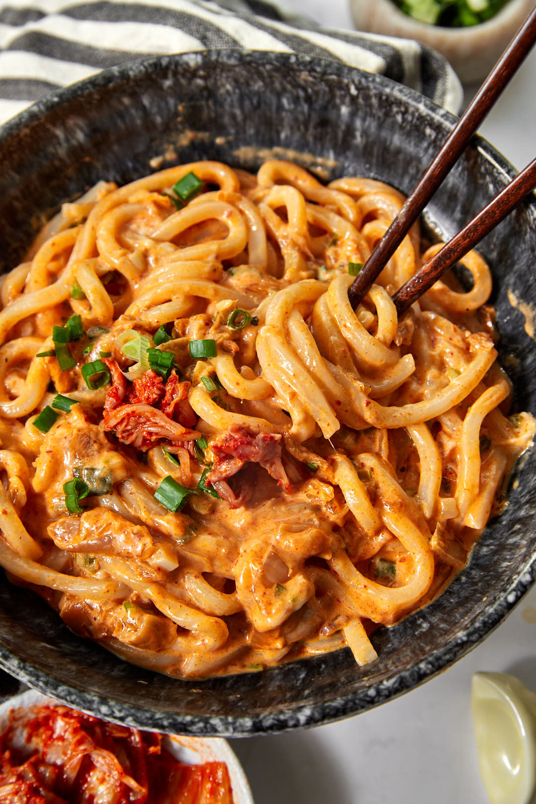 a black bowl full of of kimchi udon noodles  garnished with green onions, with chopsticks sticking into the bowl.