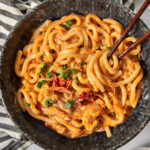 a black bowl full of of kimchi udon noodles garnished with green onions, with chopsticks sticking into the bowl.