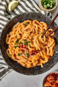 a black bowl full of of kimchi udon noodles garnished with green onions, with chopsticks sticking into the bowl.