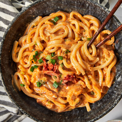 a black bowl full of of kimchi udon noodles garnished with green onions, with chopsticks sticking into the bowl.