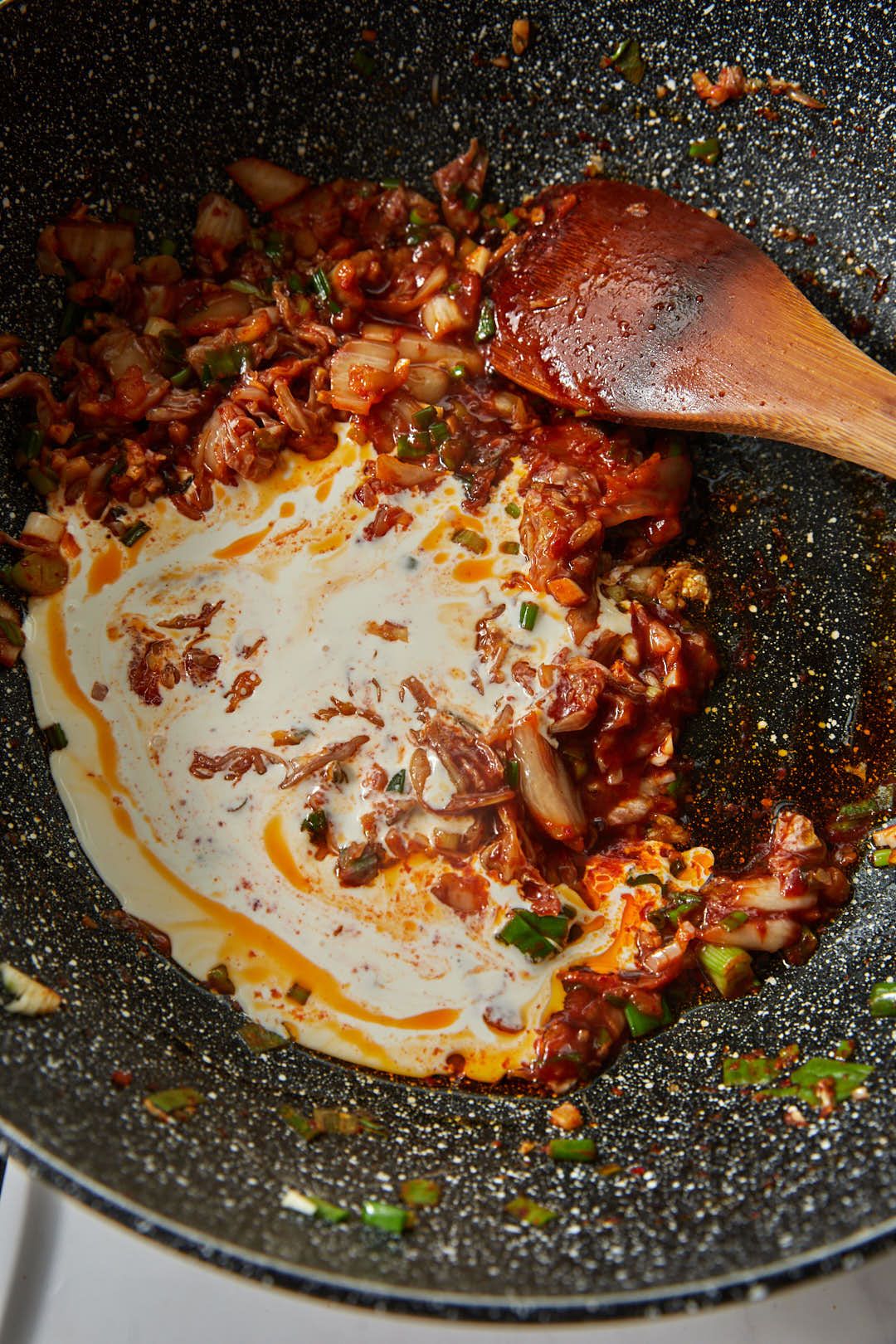 cream being added to gochujang and chopped kimchi in a large skillet with a wooden spoon.