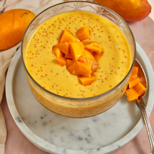 photo of mango sago in clear trifle dish on a marble plate with ripe mangoes in the background.