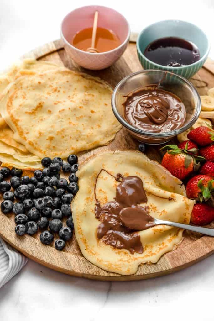 nutella crepe being assembled on a wooden board with strawberries, berries and bananas