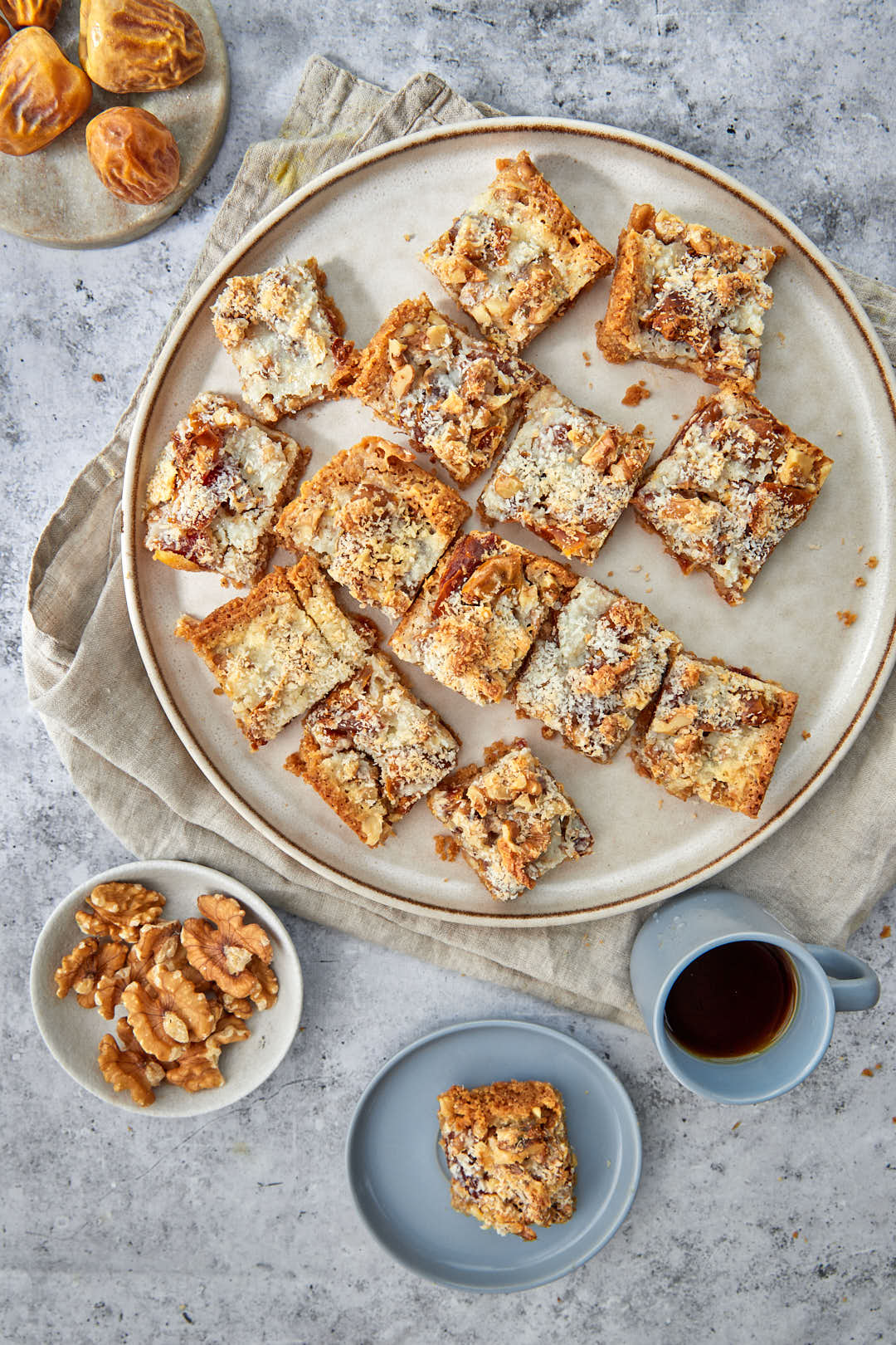 a plate of date coconut squares with a cup of coffee and some walnuts on the side.