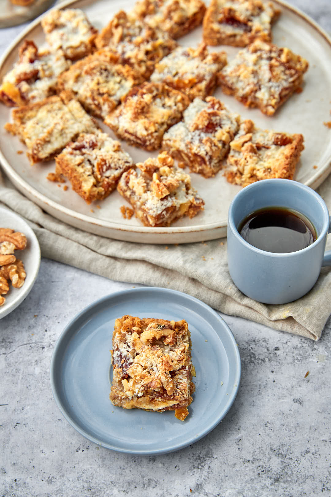 a date square on a blue plate with a cup of coffee and a larger plate of date squares in the background.
