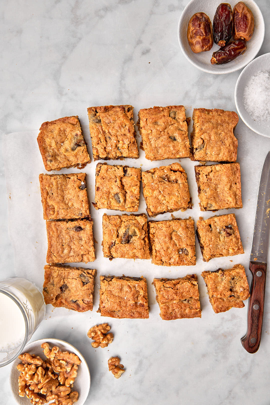 date blondies cut into squares on a white marble background, with walnuts and a glass of milk on the side.