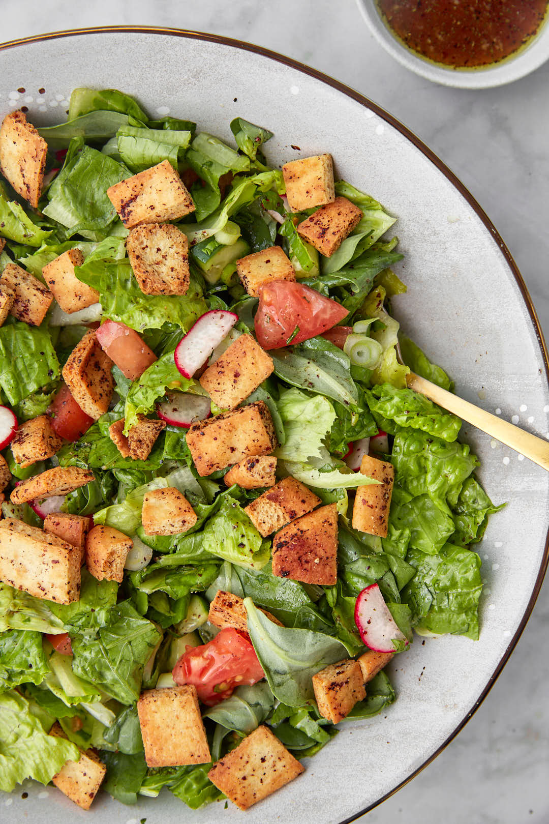 a big bowl of fattouche with a serving spoon in it and extra dressign on the side.