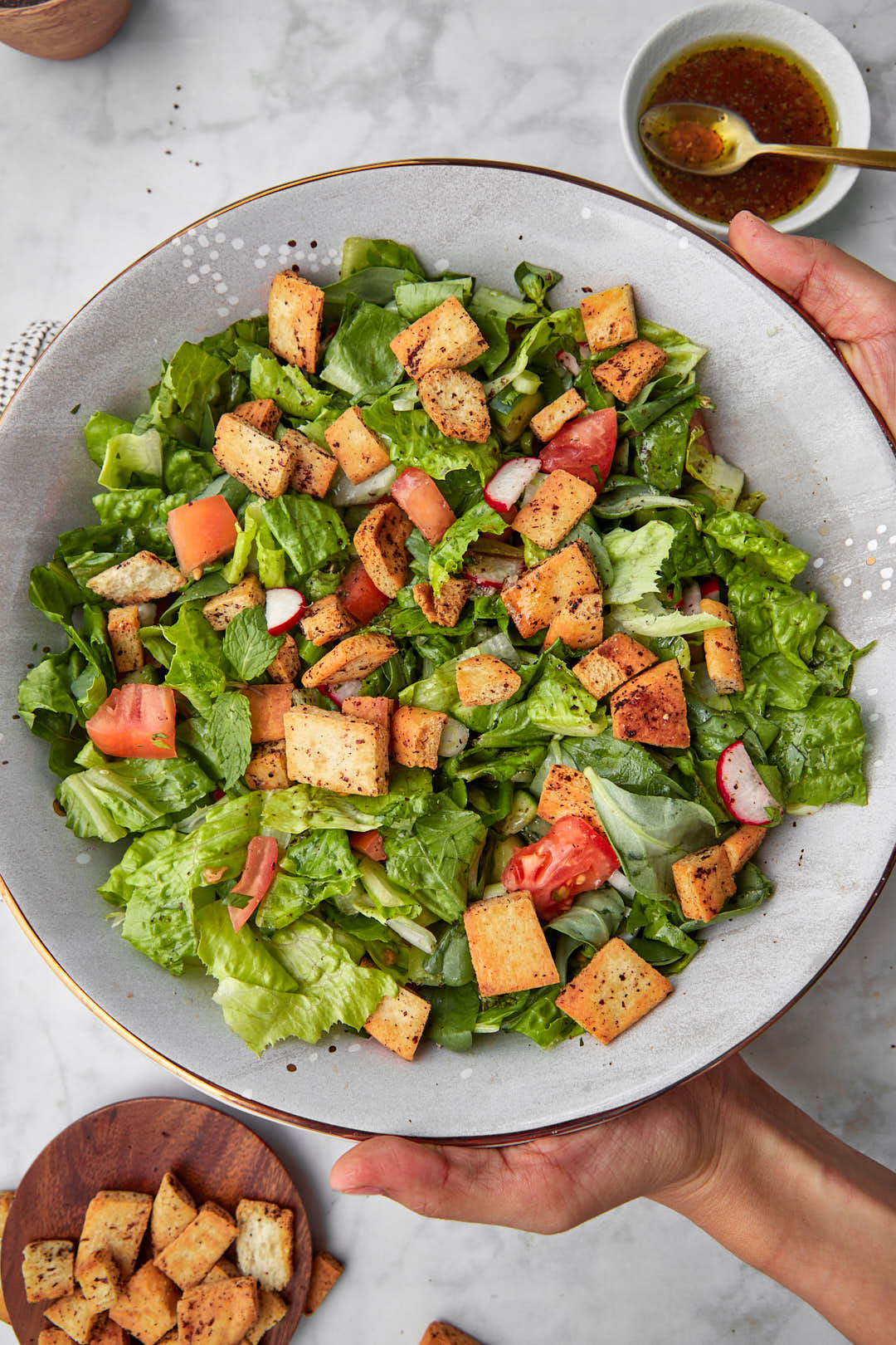 two hands holding a finished plate of fattouche salad with dressing in the background.