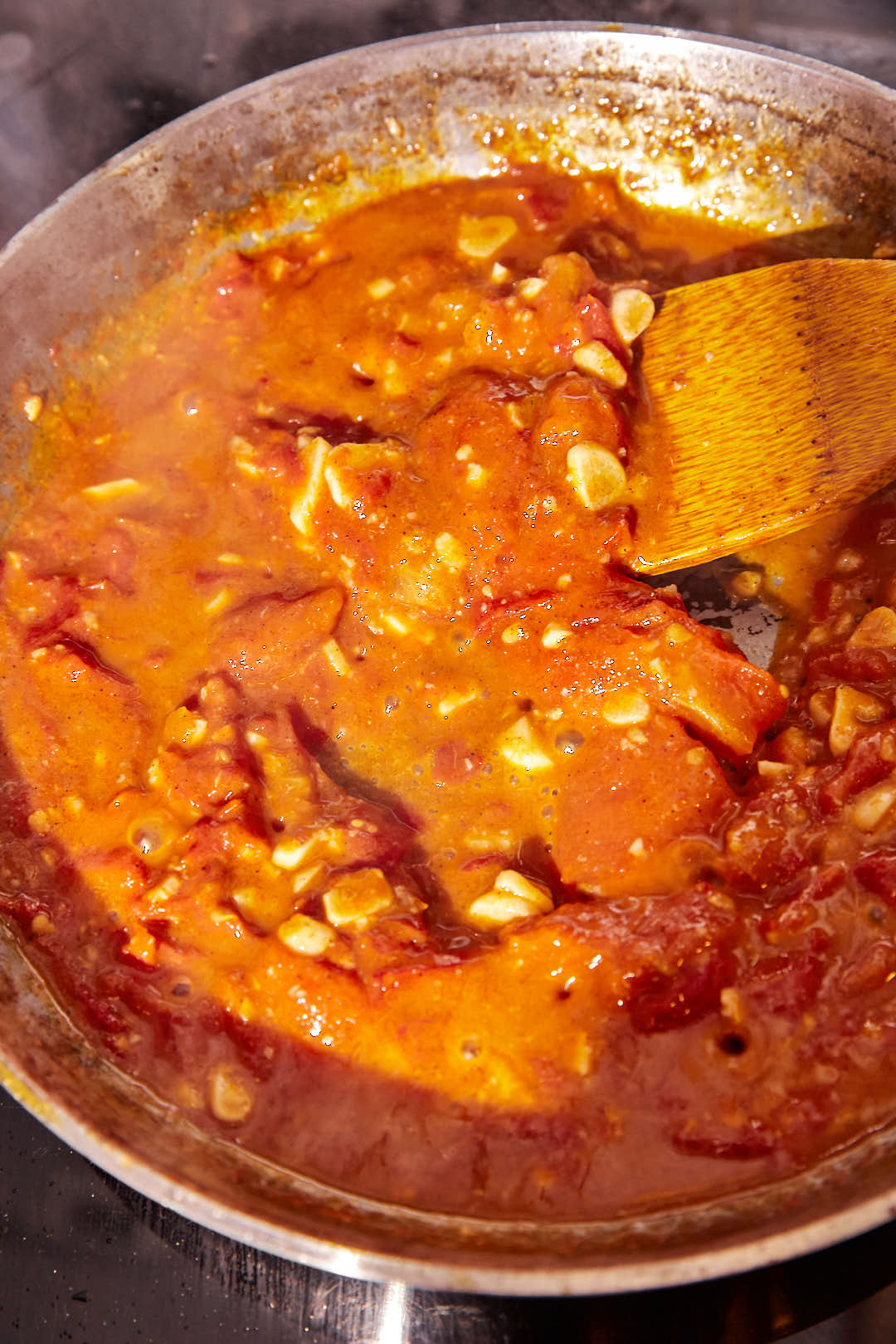 tomatoes breaking down in a skillet with garlic cloves with a wooden spoon.