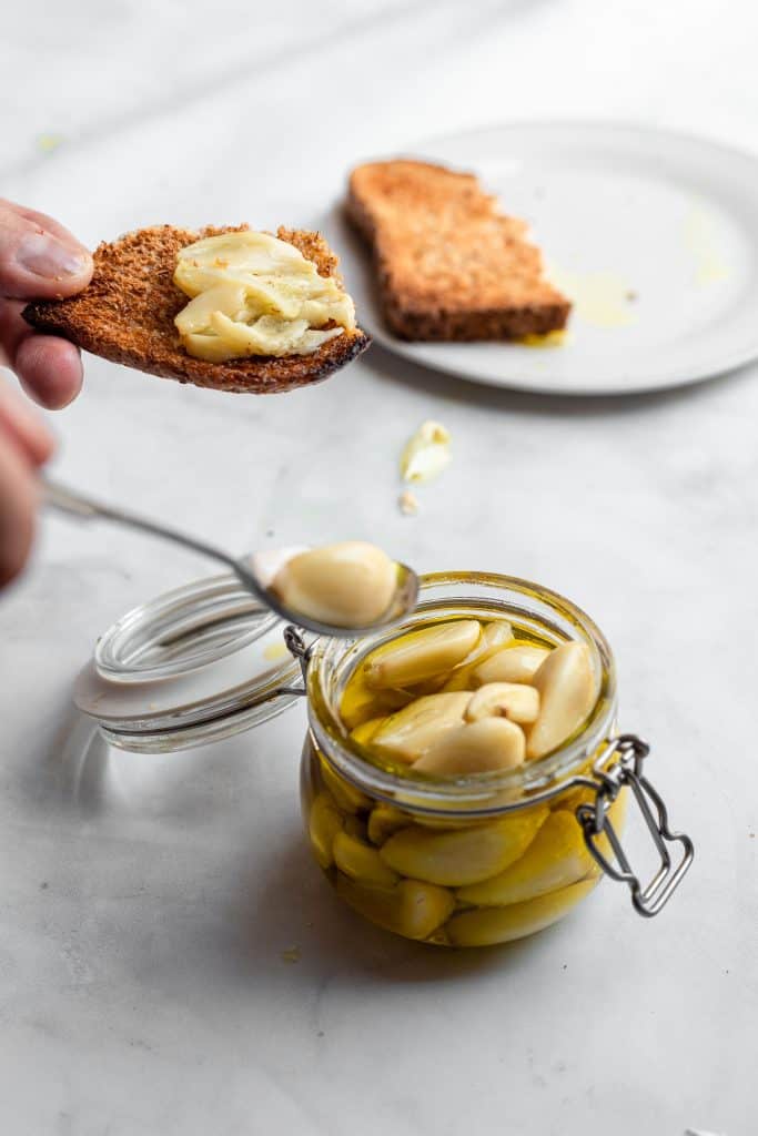 Slice of bread with garlic confit spread on it and more garlic being spooned out of a jar