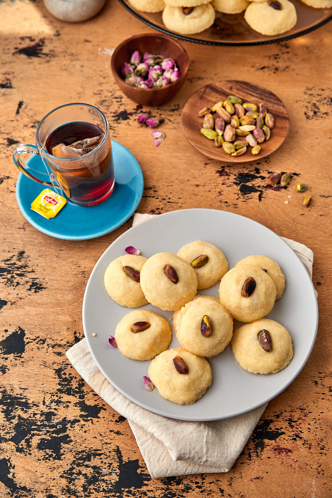 a plate of ghraybeh cookies with a cup of tea in the background and a plate of pistachios in the background.