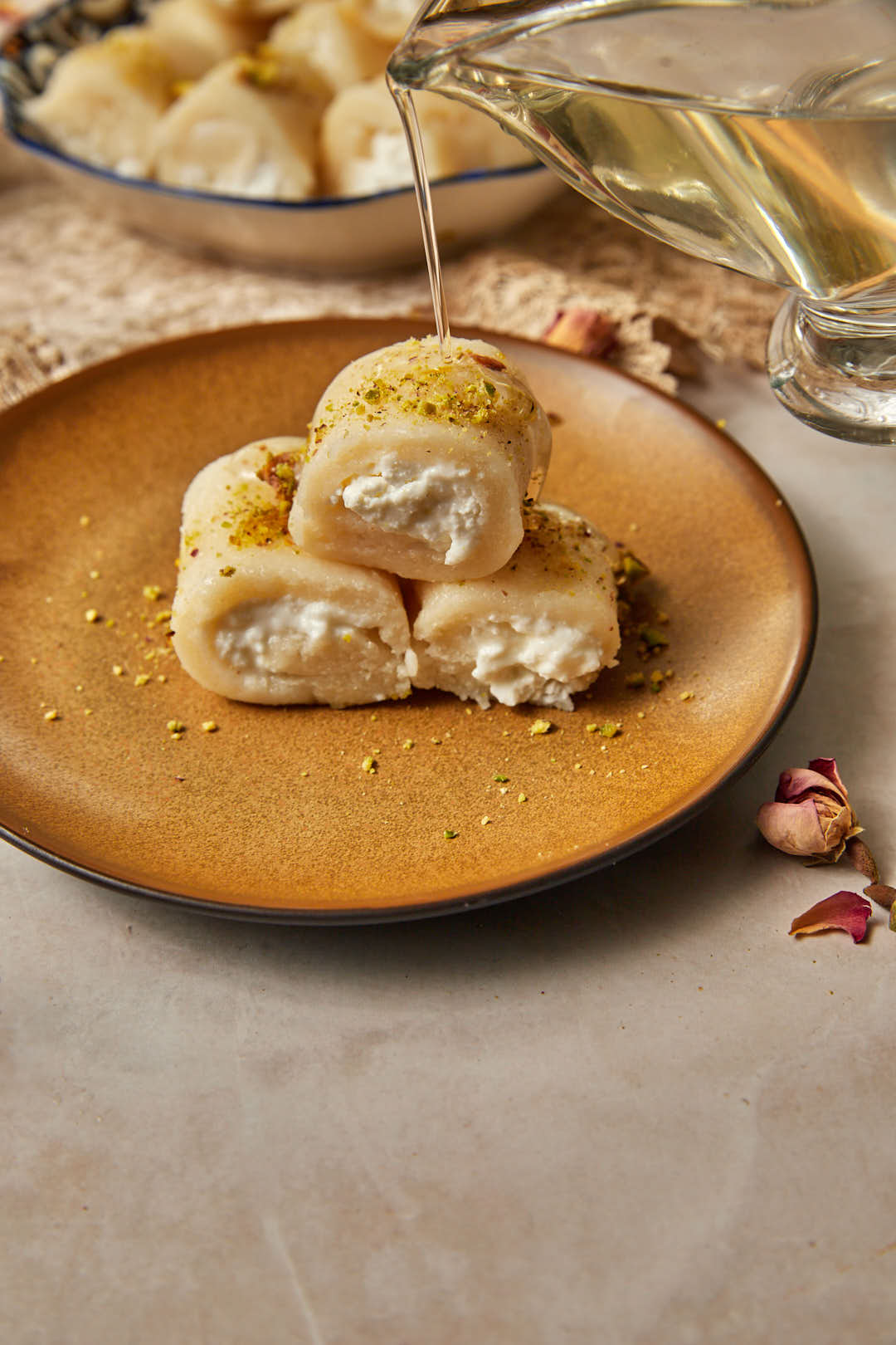 sugar syrup being drizzled onto three rolls of halawet el jibn on a brown plate.