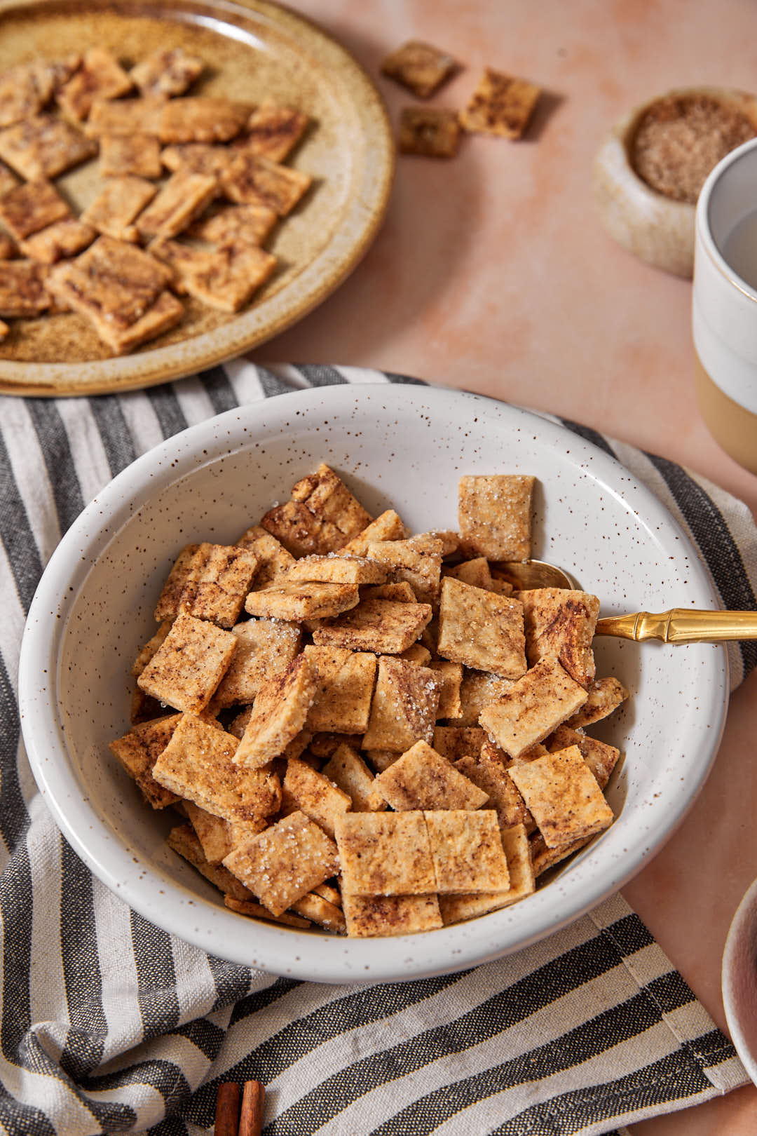 a bowl full of healthy cinnamon toast crunch with a spoon in it, without milk. A plate of more cinnamon toast crunch in the background.