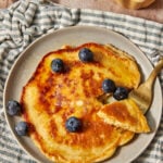 a large cottage cheese pancake with blueberries scattered on top, on a blue and white striped linen towel