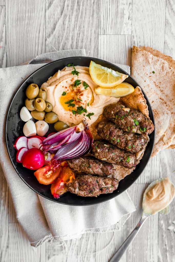 a plate of cooked kafta with hummus and veggies and olives with pita bread on the side